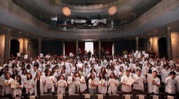 VÁRIOS JOVENS USANDO UNIFORME E AVENTAL  BRANCO NA PLATEIA DE TEATRO. tODOS SEGURAM SEUS CERTIFICADOS