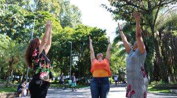 Três pessoas levantam a mão para o alto, no meio de uma alameda do Jardim Botânico. Elas parecem alongar o corpo. #Pracegover