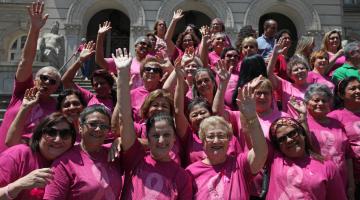 Mulheres vestidas com camisetas cor de rosa posam para foto em frente à Prefeitura. Elas estão com os braços para o alto.#Pracegover