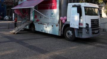 Caminhão estacionado em praça. O veículo tem  uma tenda na frente e uma rampa de acesso ao seu interior. #Pracegover