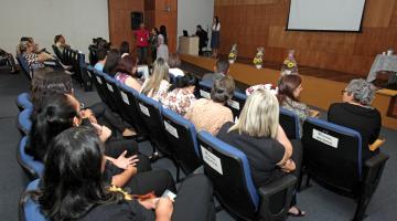 Pessoas sentadas em cadeiras de auditório prestam atenção em que está no palco, ao fundo. #Pracegover