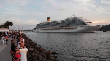 navio está saindo do porto de santos e pedestres estão próximos à mureta da ponta da praia para avistá-lo. #paratodosverem 