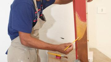 Homem está pintando peça de madeira. Há potes de tinta e pinceis sobre a mesa. #Pracegover