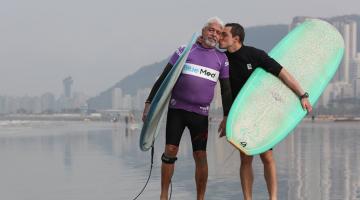 pai e filho estão na beira da praia, ambos segurando pranchas. Filho abraça e beija o rosto do pai. #paratodosvere