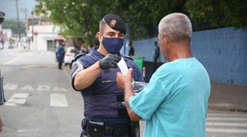 #pracegover Com máscara no rosto, guarda municipal entrega máscara descartável a idoso em calçada de avenida