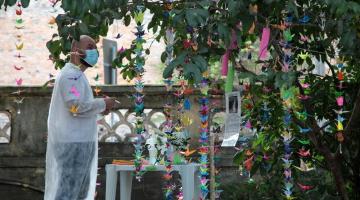 Enfermeiro com caneta e papel na mão observa árvore cheia de origamis coloridos. #paratodosverem