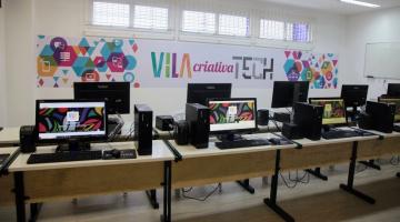 sala com diversos computadores sobre escrivaninhas. Ao fundo na parece há uma arte e os dizeres Vila Criativa Tech. #paratodosverem 