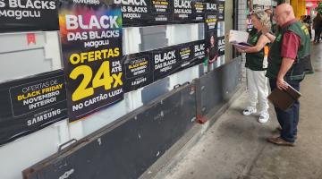 dois agentes uniformizados do procon estão na rua diante da vitrine de uma loja com cartazes sobre a black friday. #paratodosverem 