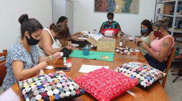 seis mulheres sentadas costurando sobre uma mesa , almofadas sobre a mesa #paratodosverem