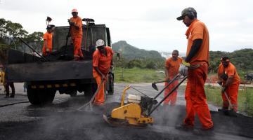 Homens uniformizados aplicam massa e nivelam solo com pás e máquina. Há um caminhão atrás dos trabalhadores. #Pracegover