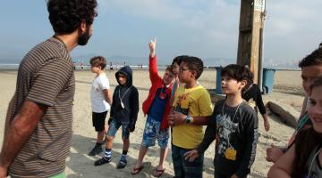 crianças estão na praia com professor e uma delas levanta o braço#pracegover 