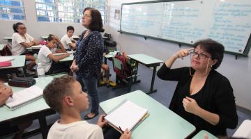 Em primeiro plano, à direita, intérprete faz liguagem de sinais para aluno que está à sua frente. Atrás, a professora dá aula para os alunos. #Pracegover