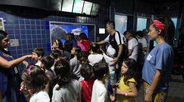 Muitas crianças estão passeando em corredor do Aquário. Elas estão conversando e observando os tanques. #Pracegover