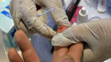 mãos com luvas seguram dedo de uma pessoa para extrair gota de sangue. #paratodosverem 