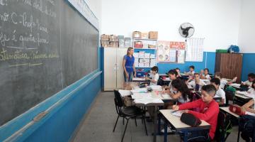 Sala de aula com professora ao fundo. A lousa está ao lado esquerdo da imagem. Os alunos estão sentados em suas carteiras. #Pracegover