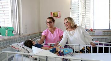 Criança está em maca de hospital. Duas mulheres estão ao seu lado e fazem carinho na criança. #Pracegover