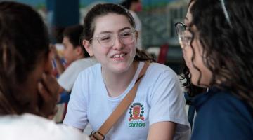 Bruna está sentada, de frente para a foto, conversando com amigas que aparecem de costas para a foto. #paratodosverem