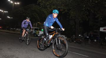 Ciclistas pedalam em avenida durante a madrugada. Na imagem aparecem dois. Eles usam uniforme e capacete. #Paratodosverem