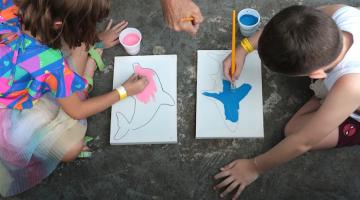 foto tirada do alto mostra duas crianças frente a frente pintando telas. A silhueta de animais marinhos está pronta. Elas pintam sobre a área delimitada.  #paratodosverem 