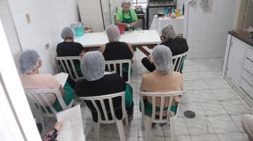 mulheres sentadas de costas para a foto, todas usando tocas, prestam atenção em aula de culinária dada por uma mulher à frente de uma mesa. #paratodosverem 