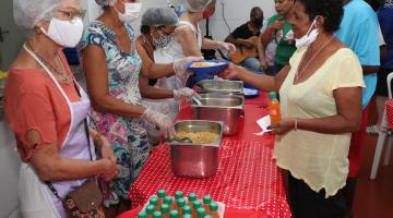 mesa com travessas fundas com comida. De um lado da mesa, pessoas servem. Do outro, são servidas. #paratodosverem