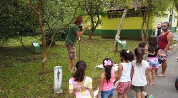 crianças estão no jardim botânico, de frente para um homemm que está à frente de uma árvore.Ele fala com todas elas. #paratodosverem