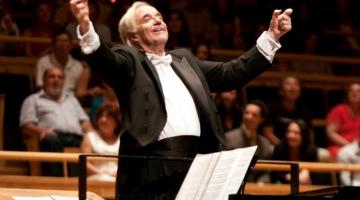 O maestro está empé, diante de partitura em estante. Seus braços estão abertos para o alto. Ele sorri. Ao fundo, o público está sentado. #paratodosverem