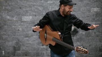 cantor está com violão pendurado ao lado do corpo. As mãos estão no ar. ele canta. #paratodosverem 