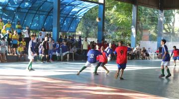 crianças estão jogando futebol em pátio. Ao fundo e e à esquerda o público está na arquibancada. #paratodosverem