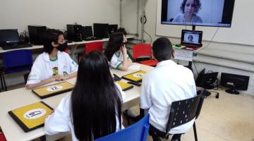 jovens estão sentados em torno de mesa assistindo apresentação em telão. #paratodosverem