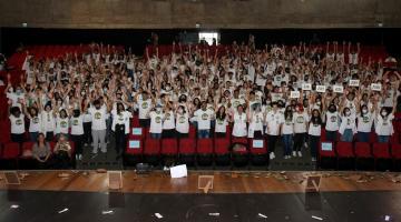 centenas de jovens uniformizados e em pé com as  mãos para o alto na plateia de teatro. #paratodosverem