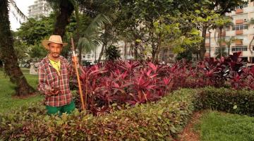 Juvenaldo posa para foto em meio a vegetação no jardim. ele usa chapéu e segura um instrumento de cabo longo. #paratodosverem
