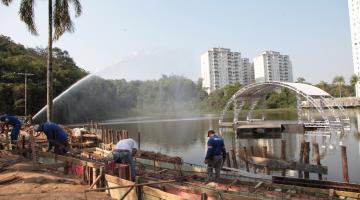 Vista geral dá área em torno da lagoa, com homens trabalhando no piso, o palco no meio da água e mais fundo, prédios do morro. #Paratodosverem