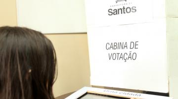 Aluna está de costas para imagem. Ela digita em uma urna eletrônica. À frente se lê em um painel: Eleição Aluno Ouvidor, Cabine de  Votação. #Paratodosverem