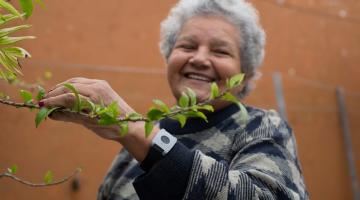 mulher está mexendo em uma planta. No braço esquerdo ela tem um aparelho, similar a um relógio. #paratodosverem