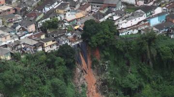 Área de morro onde houve deslizamento. Imagem feita com drone mostra o alto do morro, com moradias e uma faixa de deslizamento na encosta. Avenida passa em baixo. #Paratodosverem