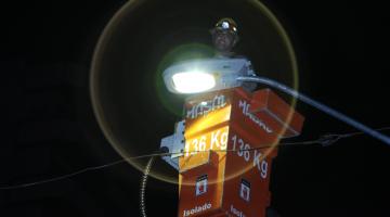 homem está em cesto no alto de poste mexendo em luminária acesa. #paratodosverem