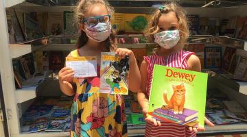 duas meninas seguram livros em frente a perua que funciona como biblioteca. #paratodosverem