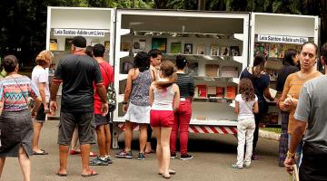 Estante com vários livros aberta em rua. Várias pessoas estão em torno. #Pracegover