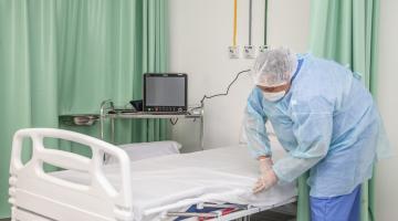 Mulher usando avental, luvas, máscara e gorro prepara leito em hospital. Ela está encaixando o lençol no colchão. Há canos de oxigênio fixados na parede atrás dela. Cortinas recolhidas fazem  o papel de biombo. #Paratodosverem