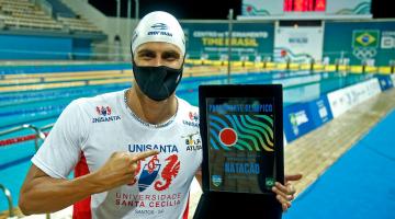 O nadador está de touca e camiseta, de costas para a piscina e segurando uma medalha que representa a classificação para a Olimpíada. #paratodosverem