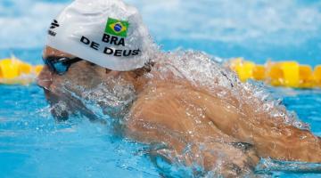 Imagem mostra o atleta nadando com toca branca com bandeira do Brasil e nome De Deus. #pracegover