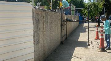 homem está sobre andaime instalando alambrado em muro. Ele está do lado de fora, na calçada. #paratodosverem 