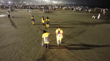 pessoas uniformizadas varrem e coletam lixo na faixa de areia à noite. #paratodosverem 