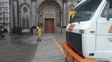 frente de caminhão de lado em primeiro plano e homem esguichando água no pixo ao fundo. Ele está à frente do portal de uma igreja. #paratodosverem