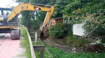 Trator com escavadeira realiza limpeza em córrego ao lado de área verde #paratodosverem