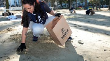 mulher agachada na areia usa luva para recolher lixo e depositá-lo num saco de papel. Ao fundo, há outras pessoas agachadas recolhendo o lixo. #paratodosverem