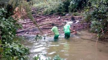 homens uniformizados estão dentro de rio com água na altura da coxa. Eles mexem em galhos que estão obstruindo trecho. Há mata nas laterais #paratodosverem