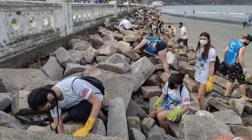 várias pessoas uniformizadas catando lixo no costão rochoso próximo à mureta na ponta da praia.Eles usam luvas e carregam sacos #paratodosverem