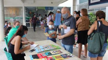 livros expostos sobre uma mesa, com duas pessoas atendendo. Uma criança olha e segura um livro ao lado de um adulto. Há uma filha de  pessoas para vacinação atrás deles. #paratodosverem